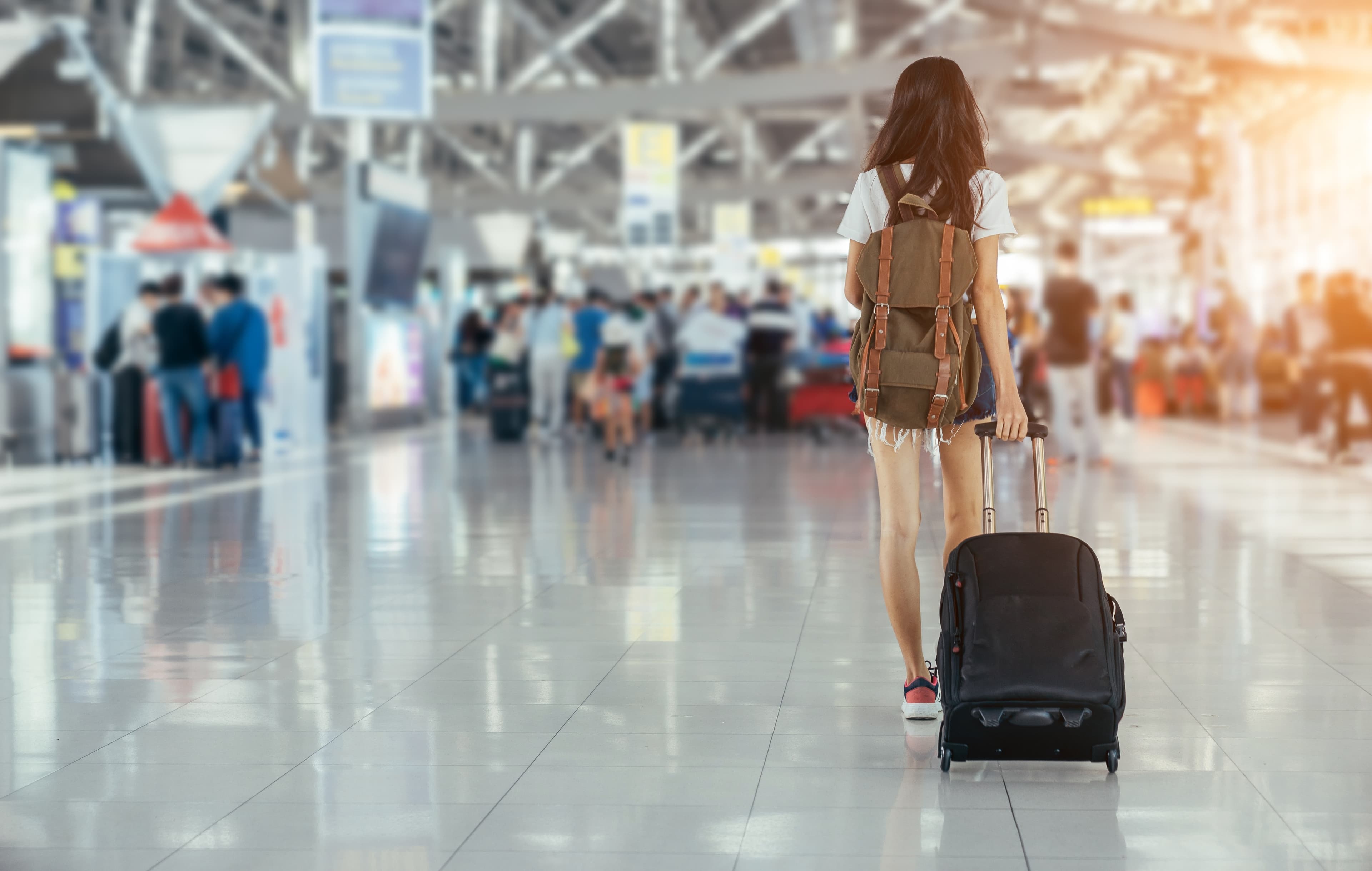 Frau mit Koffer an Flughafen. Im Hintergrund Flughafenbetrieb.