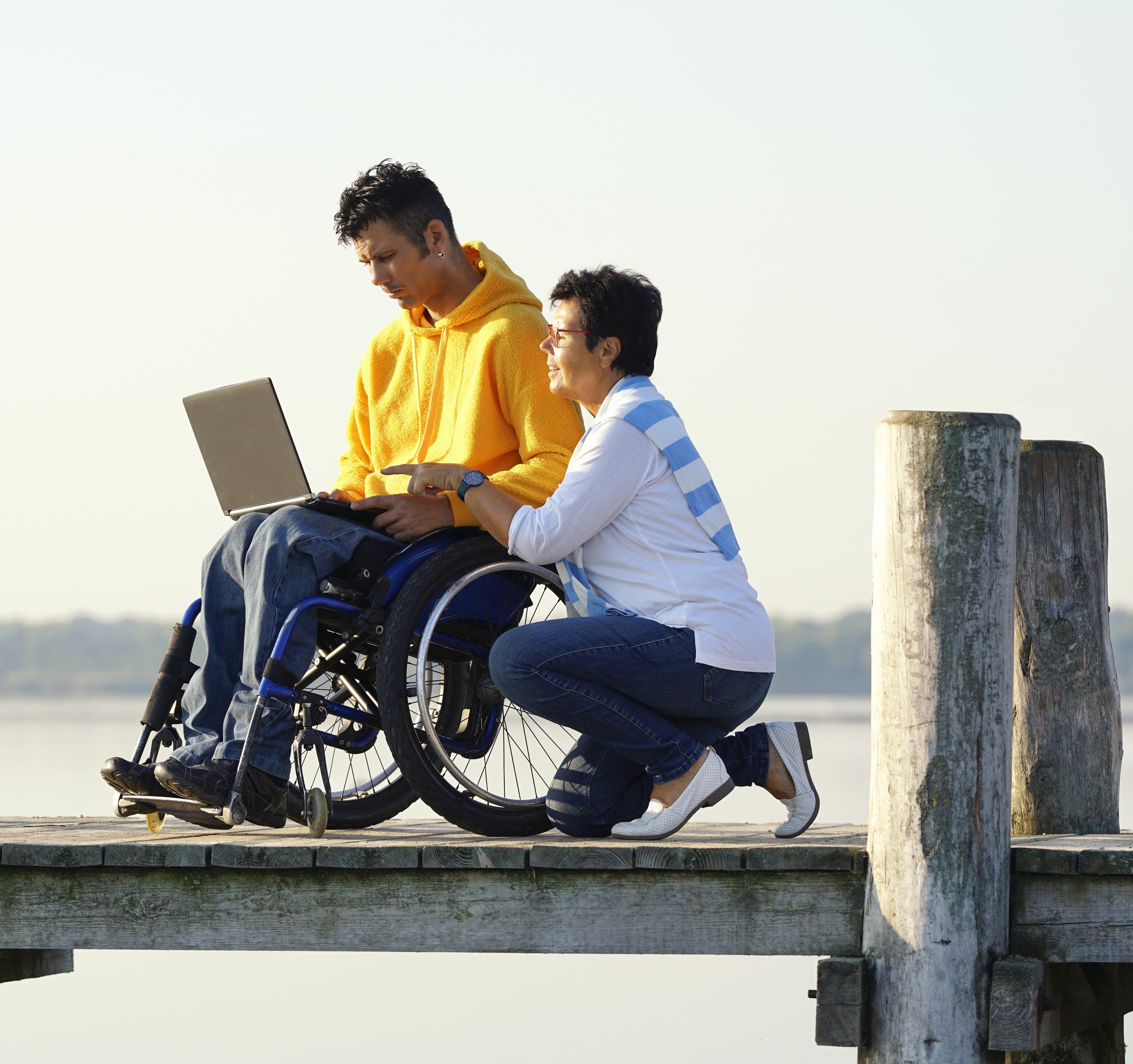 Mann in Rollstuhl am Laptop auf Steg über Wasser. Frau kniet daneben. Beide schauen auf Laptop.