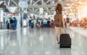 Frau mit Koffer an Flughafen. Im Hintergrund Flughafenbetrieb.